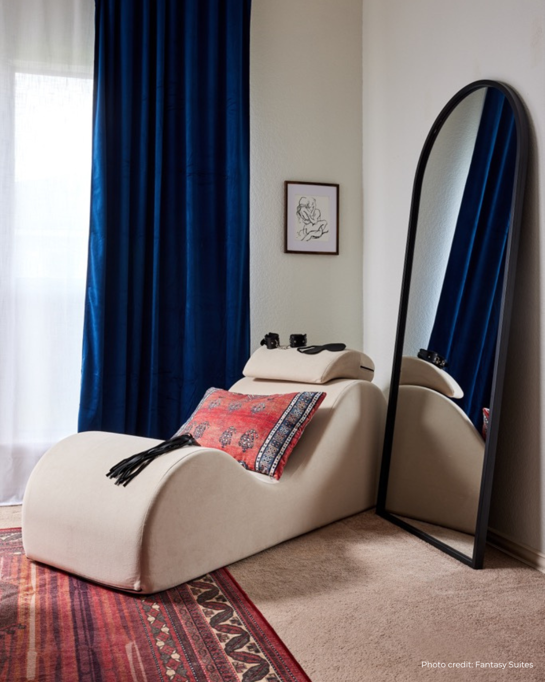 Beige sex chair with a red patterned blanket next to a standing mirror against a blue curtain.